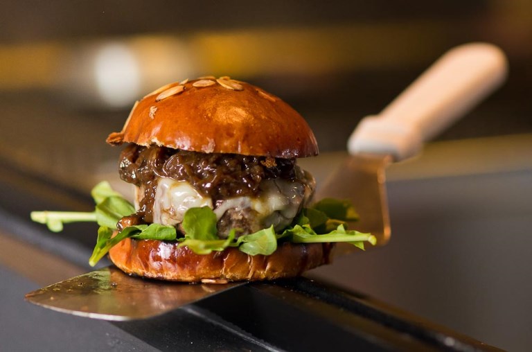 Hambúrguer da casa: pão com lâminas de amêndoa, queijo prato, cebola caramelada e rúcula Hambúrguer da casa: pão com lâminas de amêndoa, queijo prato, cebola caramelada e rúcula