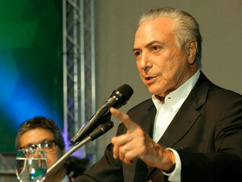 Michel Temer aparece fazendo um discurso, com o microfone preto à frente e o dedo indicador estendido