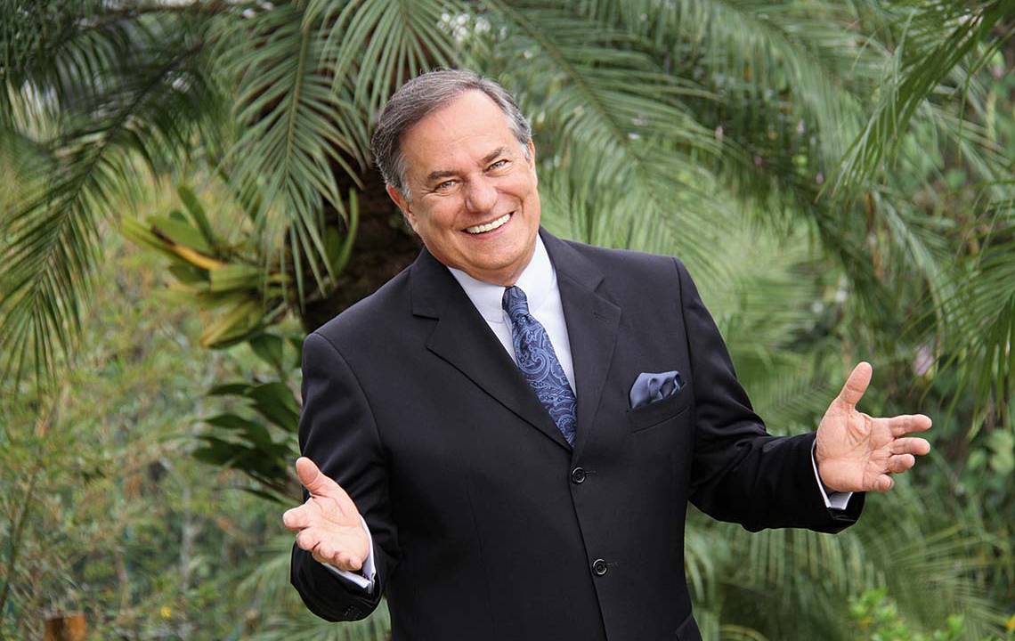 Apresentador Ronnie Von aparece de terno com as mãos estendidas, sorrindo com fundo de palmeiras.