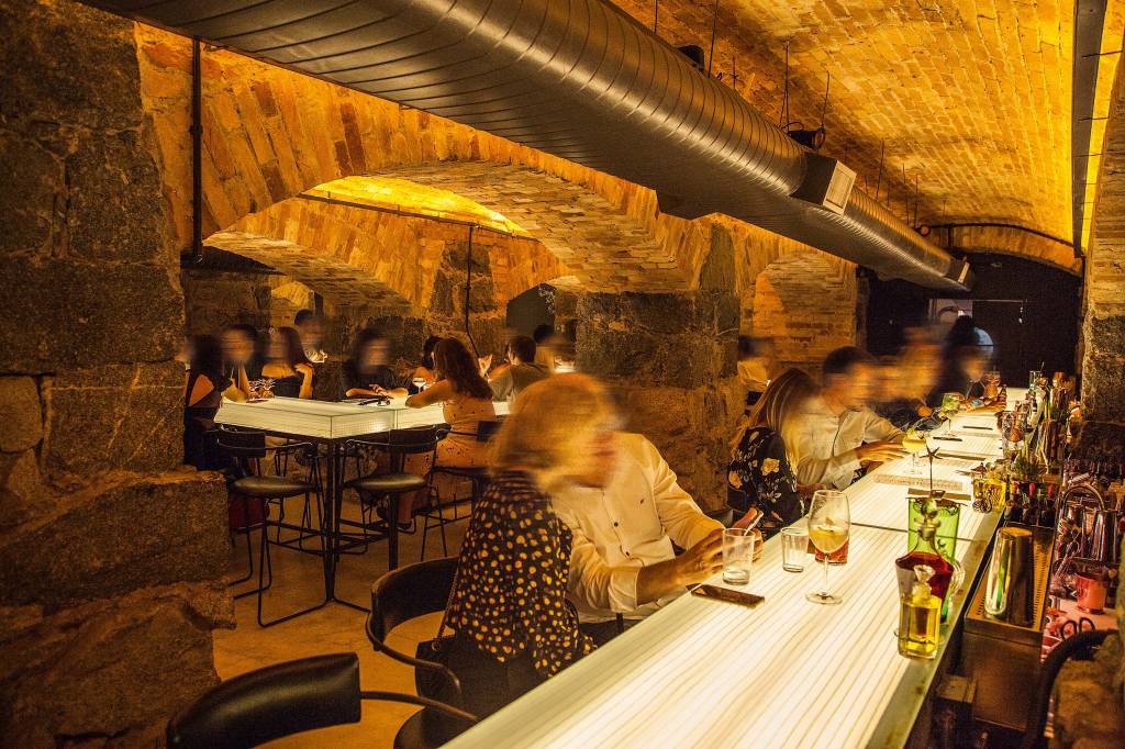 Bares pontos turísticos – Bar dos Arcos Balcão de bar iluminado com pessoa sentadas mais mesas ao fundo