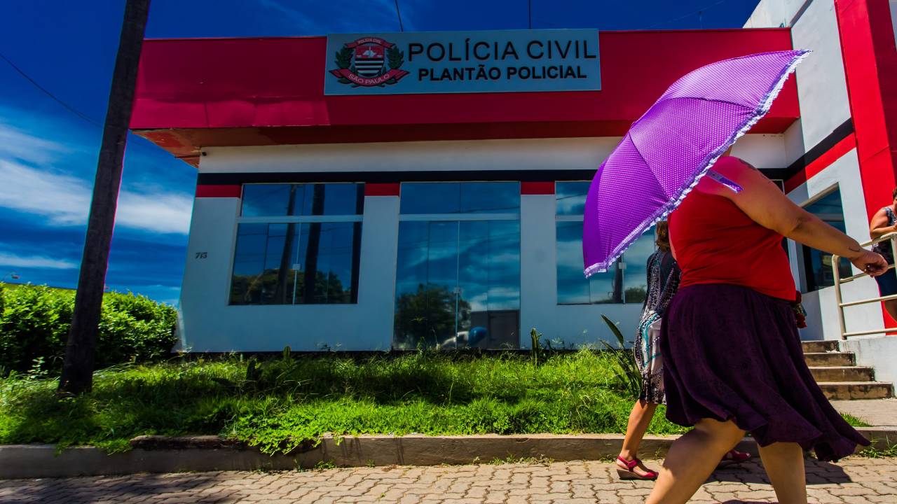 Fachada de unidade de Delegacia de Defesa da Mulher com pedestre segurando guarda-chuva rosa à direita.