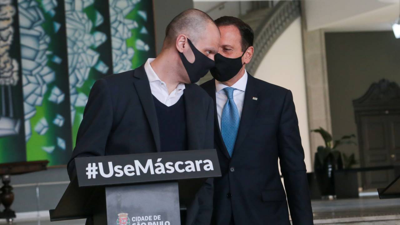 A imagem mostra Doria falando no ouvido de Covas; ambos estão de máscara