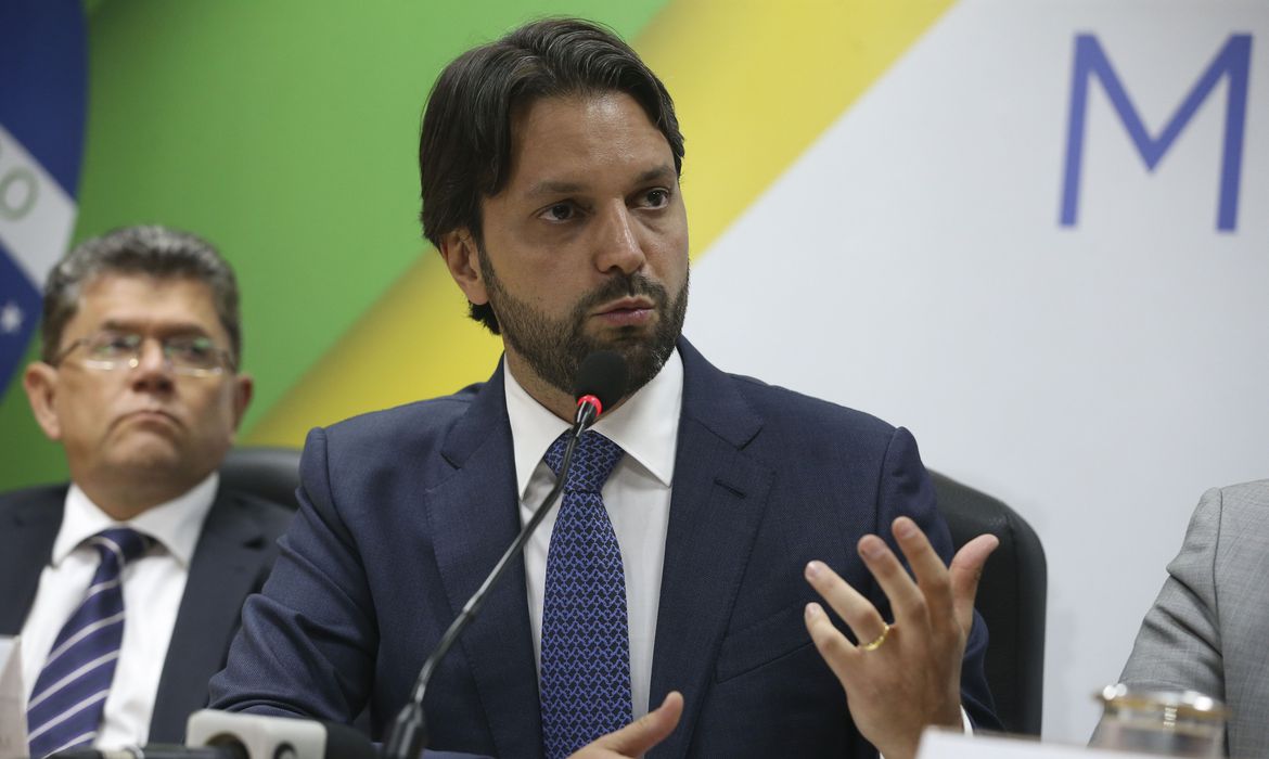 A imagem mostra Alexandre Baldy, de terno, falando em frente a microfone