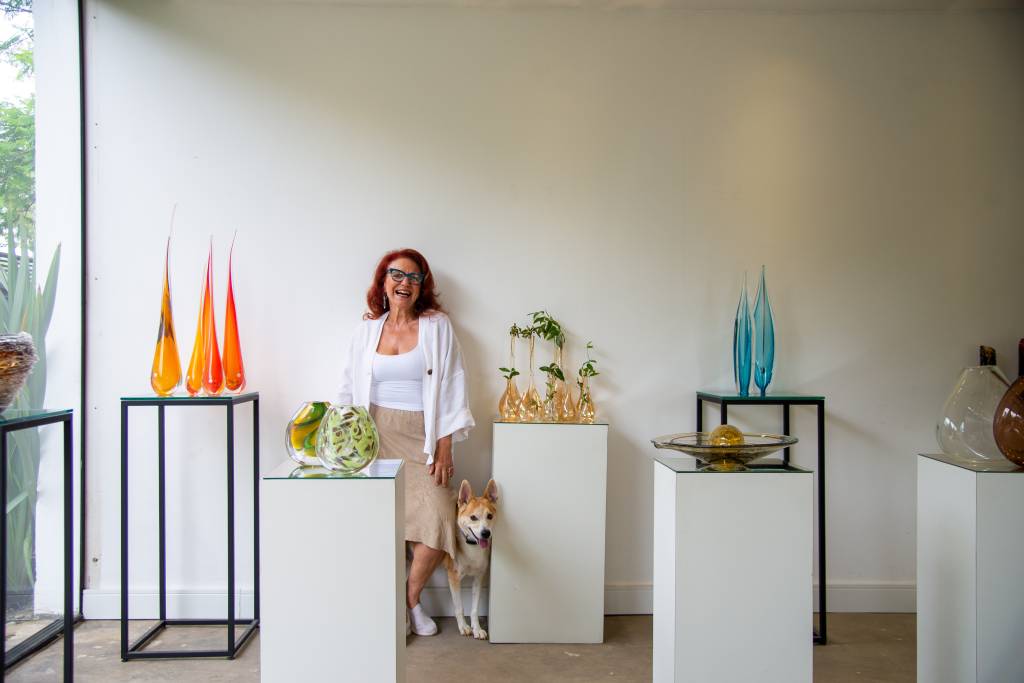 Elvira Schuartz – Espaço Zero (Rogério Pallatta) De cabelos ruivos, roupas brancas e com óculos de hastes azuis, a artista plástica Elvira Schuartz aparece sorrindo de pé e encostada na parede branca de seu ateliê, em uma área com colunas brancas que exibem peças de vidro de seu acervo nas cores laranja, azul, bege e amarelo. Ao seu lado, o cachorro da raça japonesa Shiba Inu.