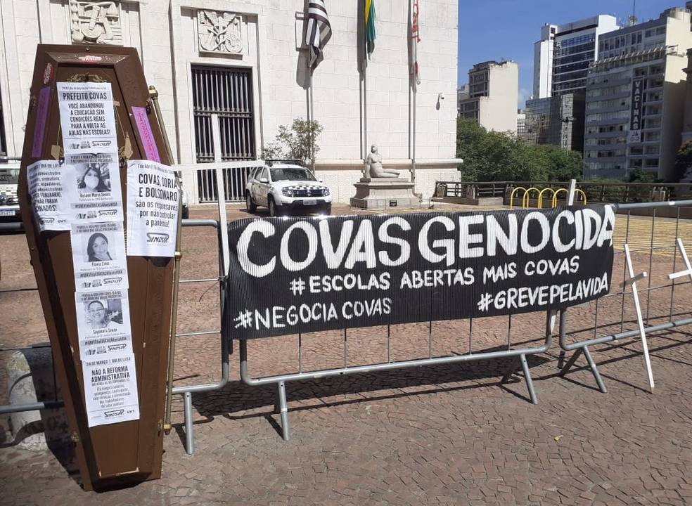 Imagem do protesto contra o retorno presencial em frente à prefeitura. Há um caixão, com fotos de professores grudados nele e também há uma faixa estendida escrito COVAS GENOCIDA ao lado, ambos apoiados em uma grade