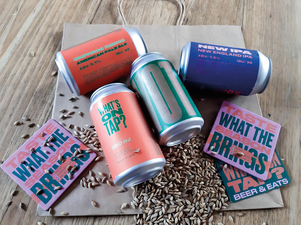 Whats on Tap – Kit com quatro cervejas enlatadas Kit com quatro opções variadas de cervejas. As latas aparecem nas cores laranja, azul e verde e estão espalhadas em cima de uma superfície de madeira.