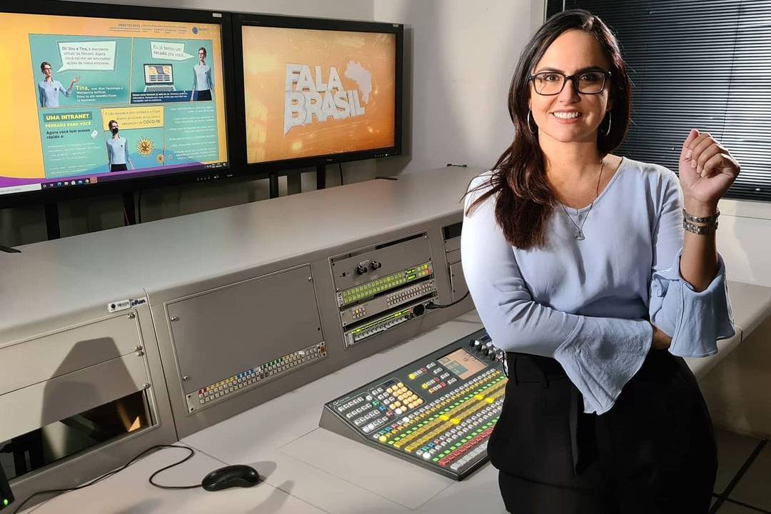 A foto mostra a jornalista Carla Cecato dentro de uma sala de operação de TV, sorrindo e posando para a câmera