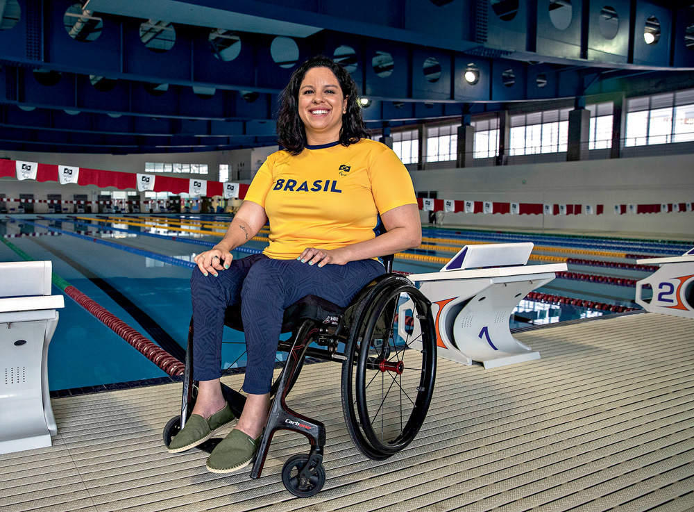 Olimpíadas-Tóquio-Paraatleta-Edenia-Garcia A fotografia mostra Edênia, de cadeira de rodas, em frente à uma piscina de natação. Ela está com a camisa amarela escrito Brasil e sorrindo para a foto.