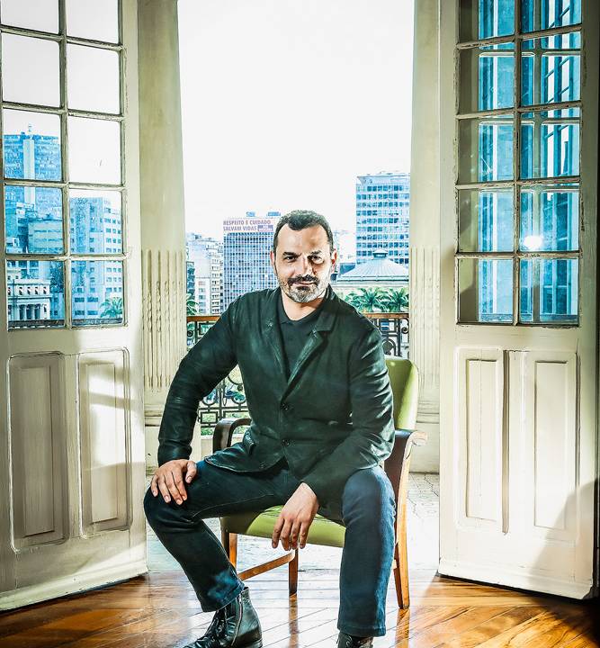 A imagem mostra Alexandre sentado em um banco em frente à uma janela da secretaria municipal da Saúde. Ele está com uma mão em um joelho e com o cotovelo apoiado no outro.