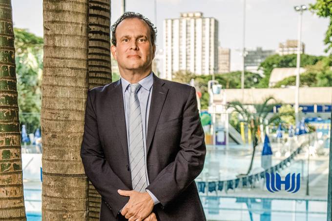 Fernando Rosenthal posa encostado em árvore com o clube Hebraica ao fundo.