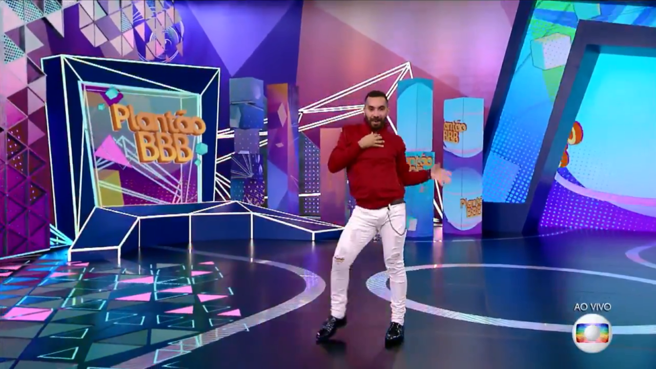 Gilberto, em pé no cenário de Plantão BBB, com uma pose com um braço no peito e outro aberto