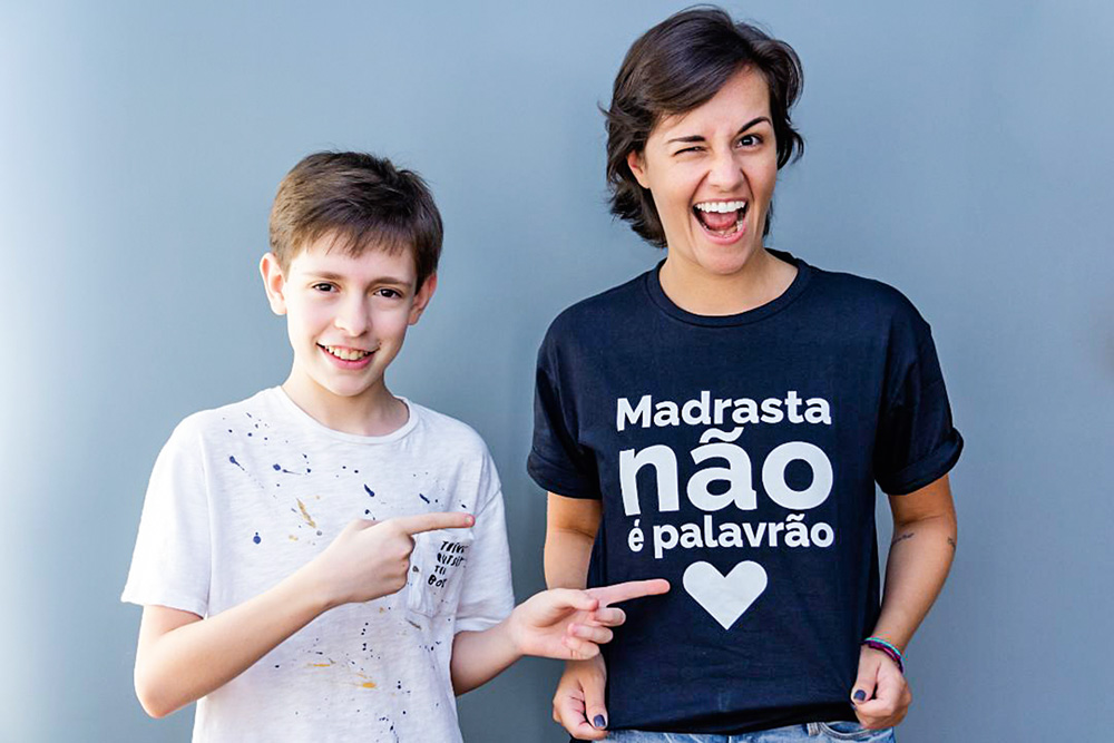 mariana destacando com as mãos a camisa posando para a foto enquanto seu enteado aponta para a mensagem da camisa