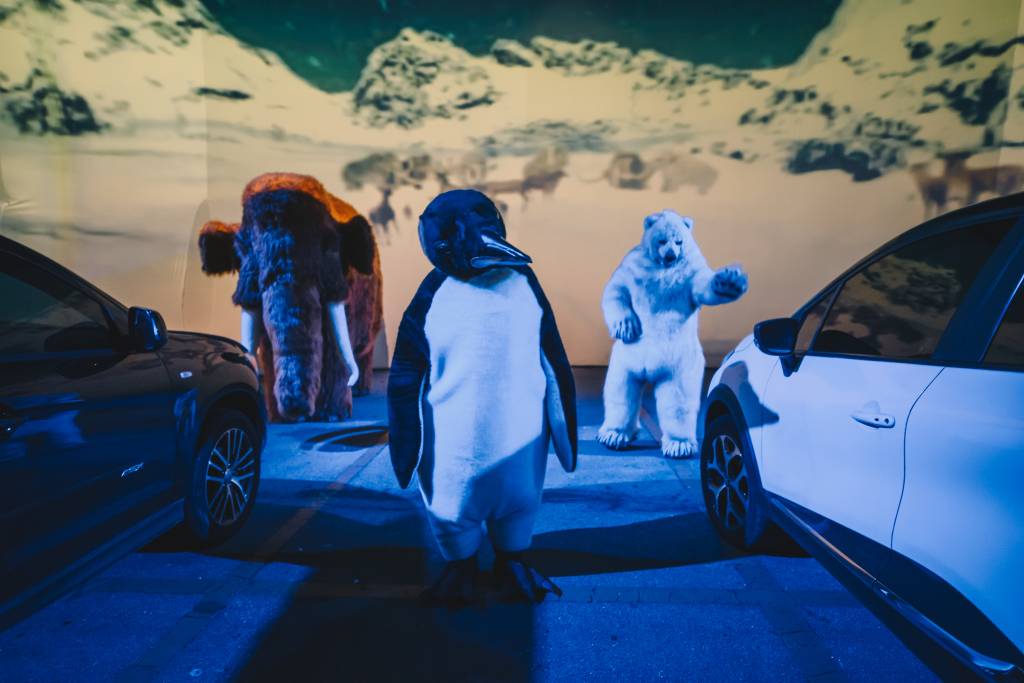 MundoAnimal_00031 Bonecos de pinguim, mamute e urso polar em frente a dois carros e telão com imagens de neve