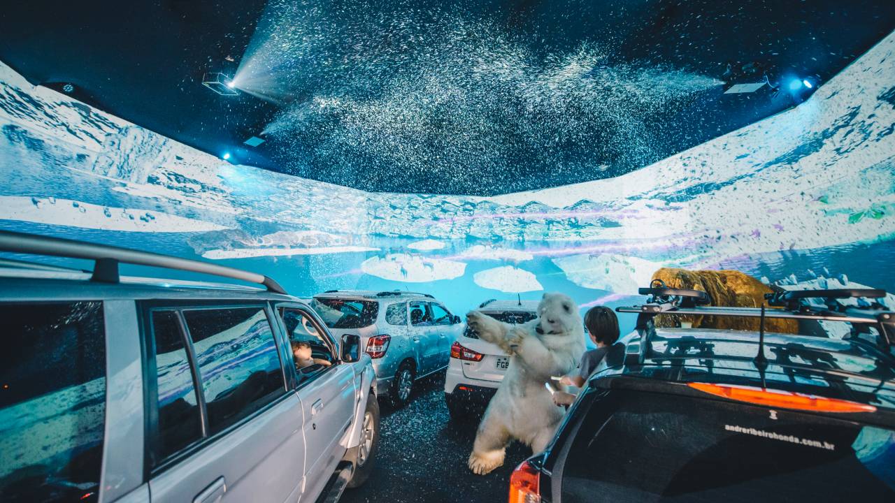 Carros parados em frente a um grande telão. Na tela, imagens de uma zona com neve e gelo. Neve artificial cai nos carros e um boneco urso polar caminha entre eles