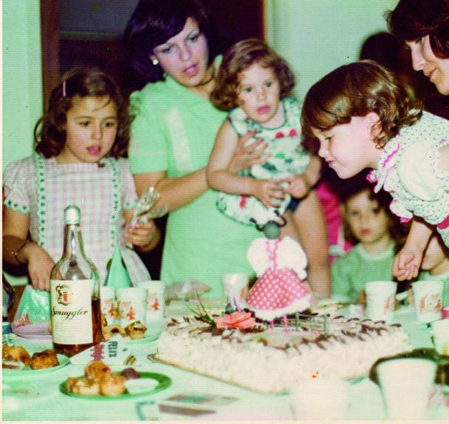 Família reunida envolta de mesa com bolo de aniversário ao centro. Criança segurada por um adulto (à esquerda) para soprar a vela do bolo.