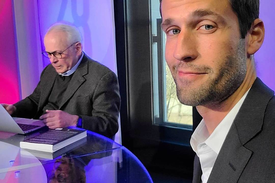 A imagem mostra uma selfie de Pedro Andrade. Ele tirou a foto de seu rosto, com um homem ao fundo, na bancada do programa Manhattan Connection