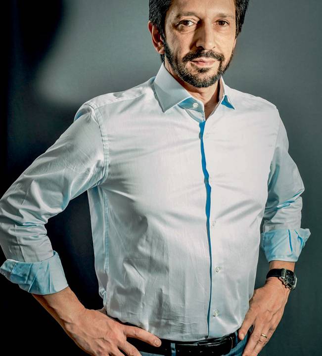A imagem é um retrato de corpo inteiro de Ricardo Nunes. Ele encara a câmera com uma expressão suave, com as mãos na cintura enquanto veste uma roupa social