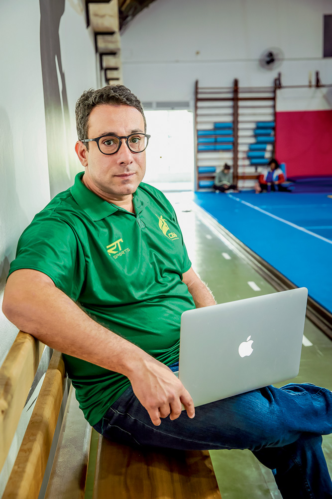 Olimpíadas-Tóquio-Handebol-Tatá A imagem mostra Tatá, com um notebook no seu colo, sentado em um banco de madeira à beira de uma quadra. Ele está com expressão serena e o uniforme da comissão técnica brasileira.