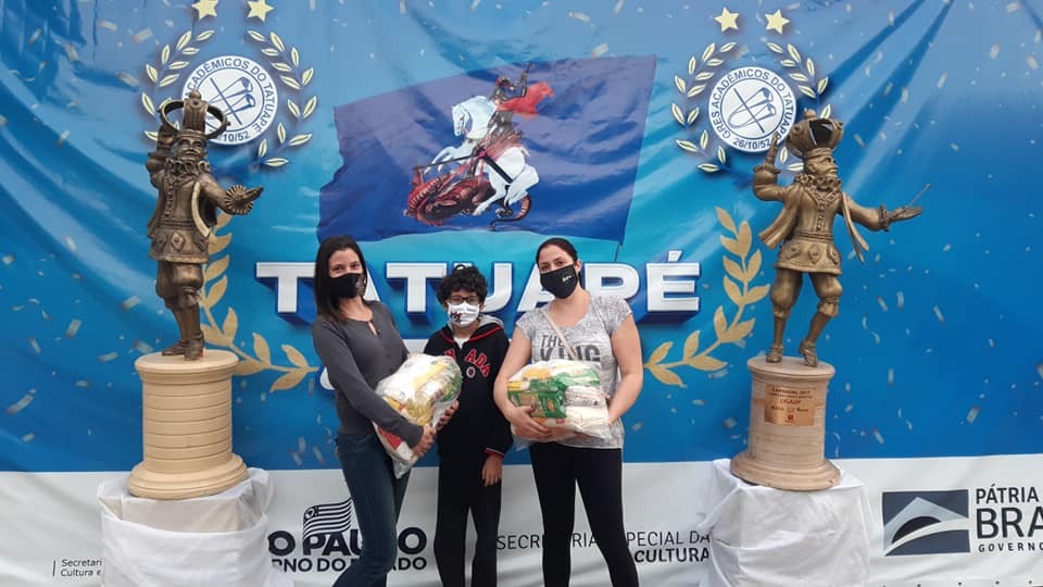 Tatuapé doações Carnaval Solidário A imagem mostra três pessoas segurando sacos de arroz, todas de máscara. Ao fundo há um bandeirão com escudos e o nome da Acadêmicos da Tatuapé