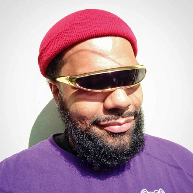 foto.jpg Homem negro com barba e touca vermelha usa um óculos metálico sem separação para cada olho