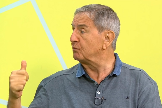 Imagem mostra Januário de Oliveira durante entrevista, olhando para a esquerda, apontando o dedão para cima; ele usa camisa polo e está em um estúdio de TV