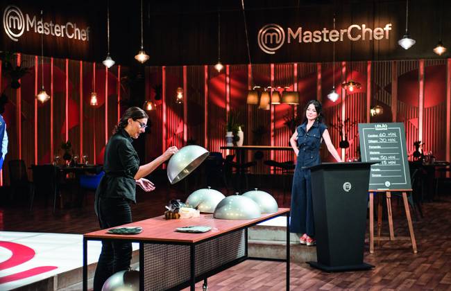 No estúdio do MasterChef. Mulher branca vestida de preto (Paola Carosella) à esquerda levantando forma esférica de prato sobre mesa de madeira. Ao fundo a apresentadora Ana Paula Padrão (mulher branca de cabelo preto) vestida de preto, encostada em púlpito do programa.