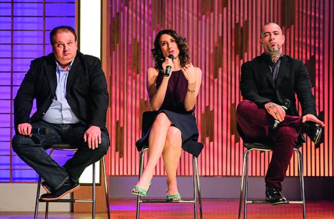 Três jurados do MasterChef sentados em banquetas em cima de palco de madeira. À esquerda, Erick Jacquin, Paola Carosella ao centro e Henrique Fogaça à direita.