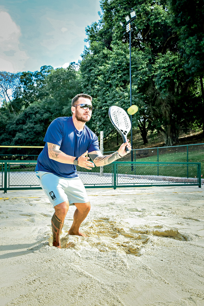 DANIEL_RECEPCAO_4.JPG A imagem mostra o professor acertando a bolinha em uma altura média. Ele está em uma quadra de beach tennis, com os joelhos flexionados para acertar a bola na altura do seu ombro.