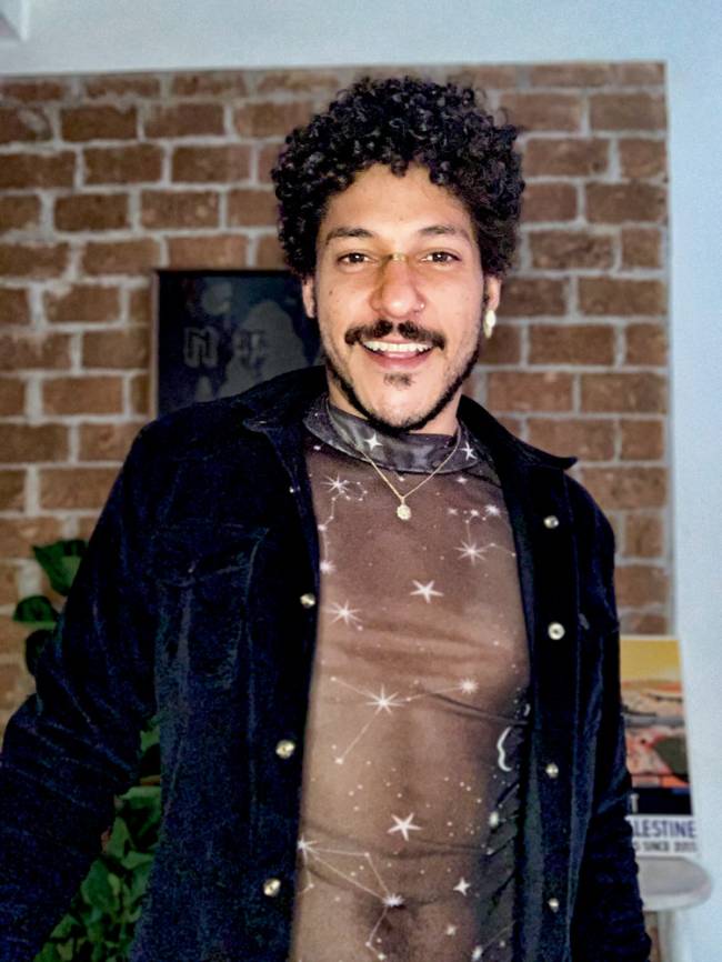 IMG-9205.jpg Homem negro com cabelos cacheados olha para a câmera sorrindo. Usa blusa transparente com estrelas