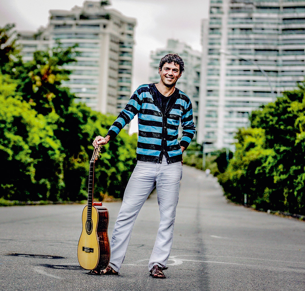 Jorge Vercillo, cantor. Um homem posa com um violão apoiado no chão ao seu lado. Ele está sorrindo em um cenário com prédios e árvores