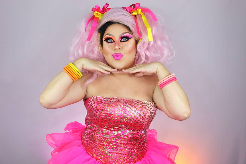 Kitana-caipira2 Drag queen posa montada com peruca, vestidos e acessórios rosa. Está com as mãos no queixo e fazendo bico com a boca