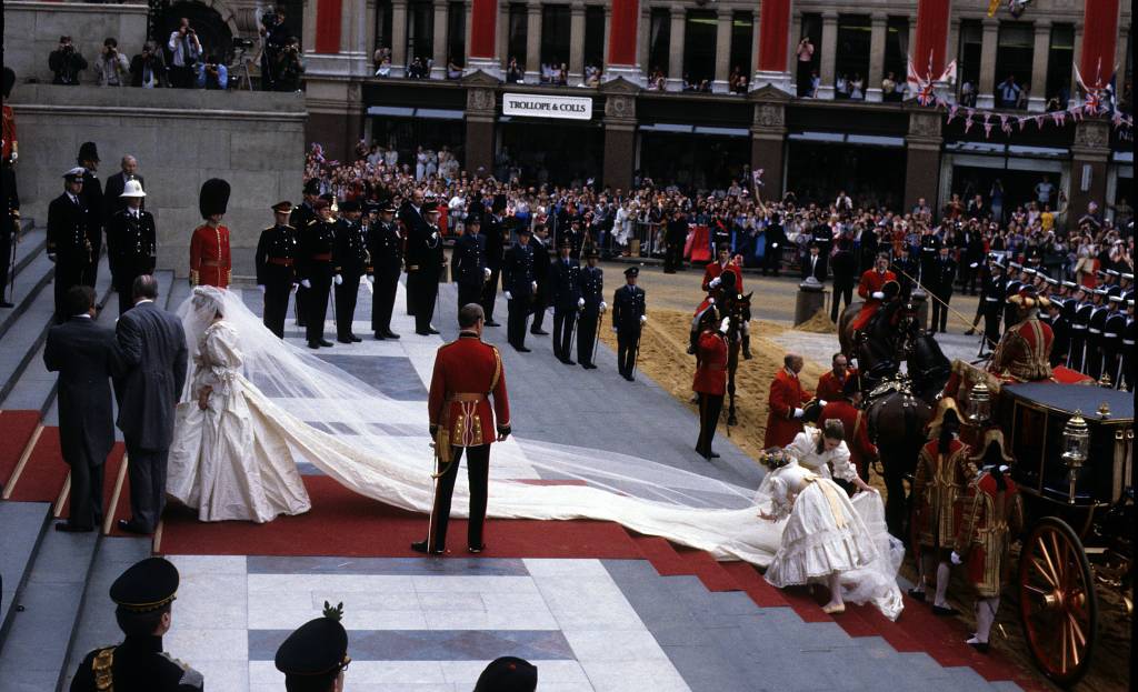40-anos-casamento-principe-charles-princesa-diana-cerimonia-chegada-Diana-igreja.jpg escadas da catedral com princesa diana pronta para entrar na catedral com seu véu segurado pelas damas de honra, próximas ao carro. diana está ao lado de seu pai, earl spencer, e há uma multidão de civis ao fundo