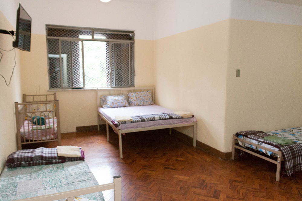 5423a0b8-c1e5-445c-8080-f2c5413d25f4 A imagem mostra um quarto no abrigo da prefeitura. É possível ver uma cama de casal, um janela, um berço e outra cama um pouco menor