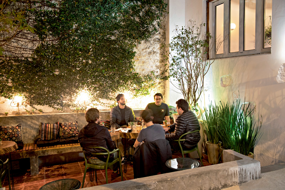 Área externa do Casca Gastrobar com cinco homens sentados ao redor de mesa de madeira cercados por vasos de flores e parede coberta por planta rasteira.