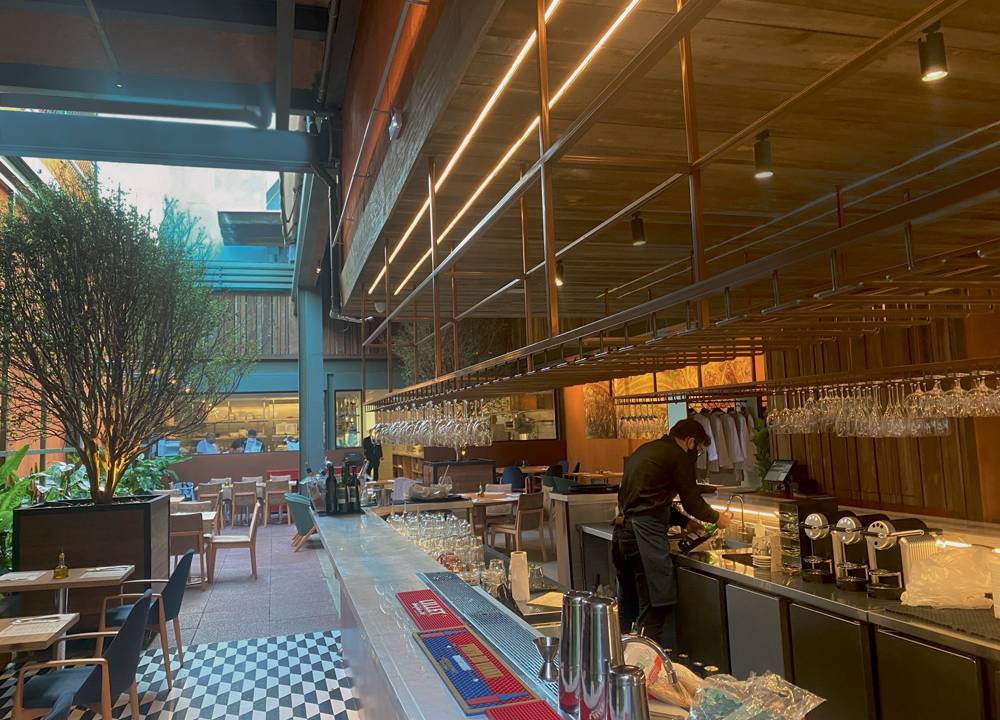 Salão da unidade de Pinheiros do Moma - Modern Mamma Osteria. Com balcão ao lado direito e mesinhas à esquerda sobre chão quadriculado.