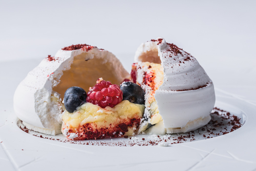 Dia dos Pais – Restaurante Fasano Em frente a um fundo branco, pavlova aberta em duas partes, com recheio de musse de mirtilo à mostra coberto por framboesa e mirtilo.