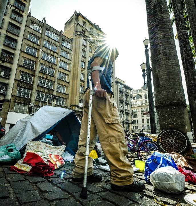 A imagem mostra uma foto, vinda do chão, observando Paulo, muleta, andando. No fundo, há uma barraca na calçada e itens espalhados pelo chão.