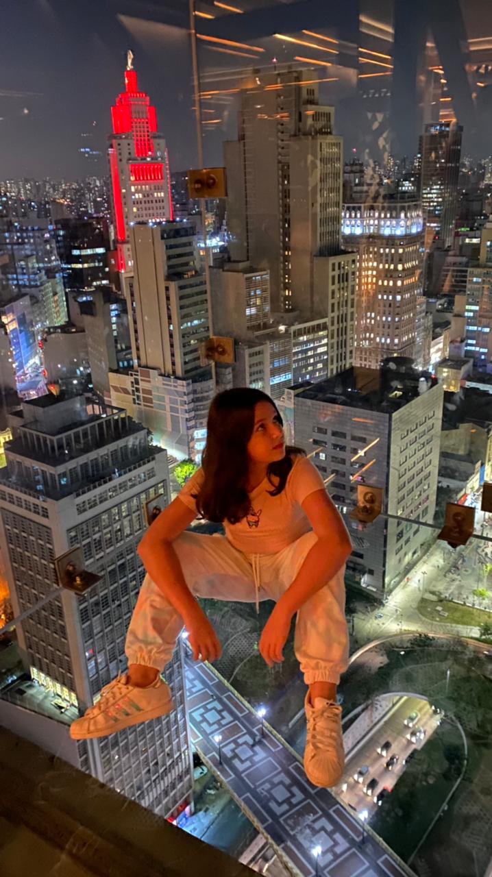 Sampa Sky (DIVULGAÇÃO) Uma menina aparece sentada no box de vidro do Sampa Sky com a vista noturna do Centro ao fundo. O Farol Santander aparece em destaque.