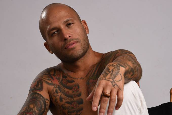 Marcelo Luna posa sem camisa sentado em cadeira de madeira. Com um dos joelhos erguidos, apoia um braço e encara a câmera. O corpo é repleto de tatuagens. Ele veste uma calça branca dobrada no tornozelo.