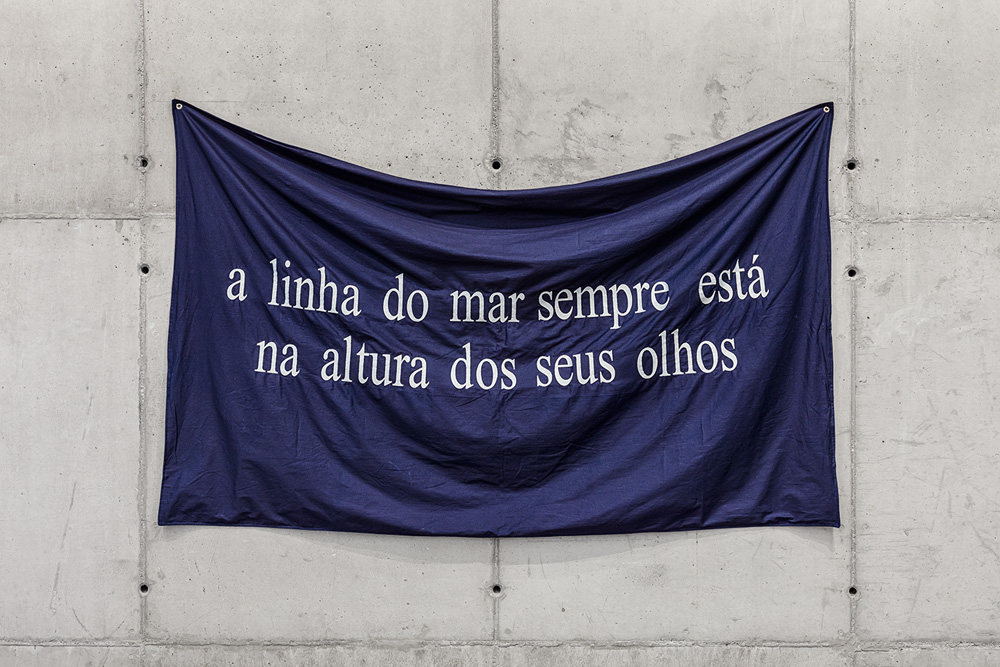 Tiago Sant_Ana_Rota de fuga, 2021_Embroidery with cotton thread on cotton fabric_109 x 188,5 cm_Photo Filipe Berndt.jpg faixa azul com a seguinte frase, em branco: a linha do mar sempre está na altura dos seus olhos