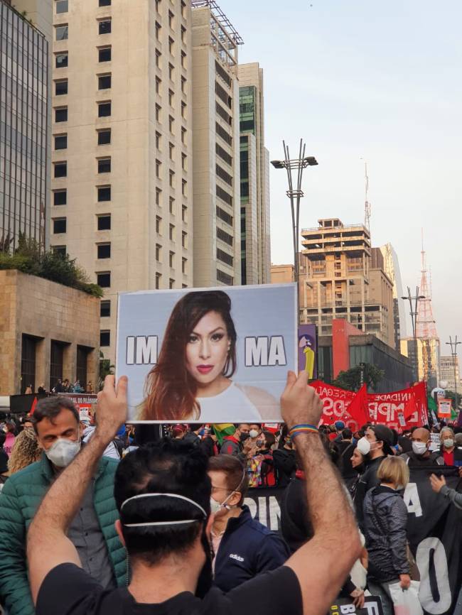 Manifestante segurando cartaz com foto da cantora Pitty em alusão ao impeatchement do presidente Jair Bolsonaro na Avenida Paulista com pôr do sol ao fundo.