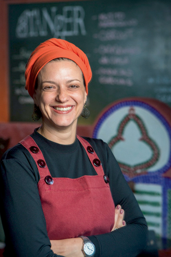 ariela-doctors-a-tal-felicidade-aromas-sabores-bem-estar-cotidiano ariela doctors está sorrindo para a foto e está utilizando um macacão cor vinho e um turbante laranja. atrás dela, desfocado, está uma lousa com coisas escritas