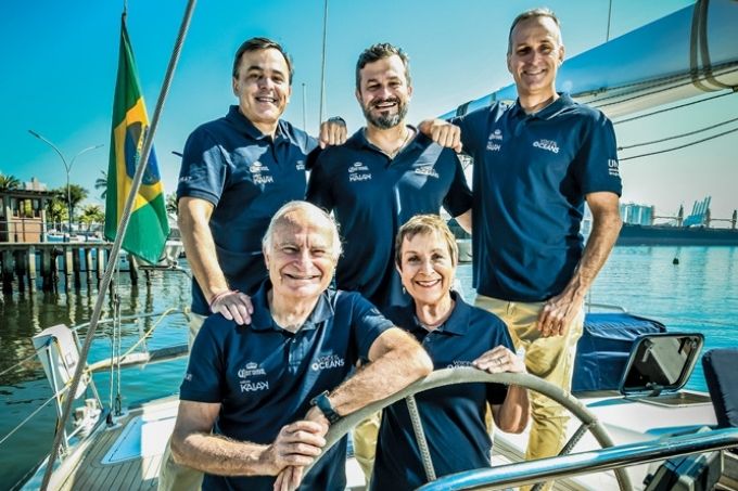 membros da família schurmann posando para a foto sorrindo à frente do volante do barco kat, com uma bandeira do Brasil ao fundo. Os membros da família estão dispostos da seguinte maneira: acima, da esquerda para a direita, estão Pierre, que está abraçando David e com mão no obro de Vilfredo, David, abraçando seus irmãos, e Wilhelm, abraçando David e com a mão abaixada. Abaixo, Vilfredo e Heloísa se apoiam no volante