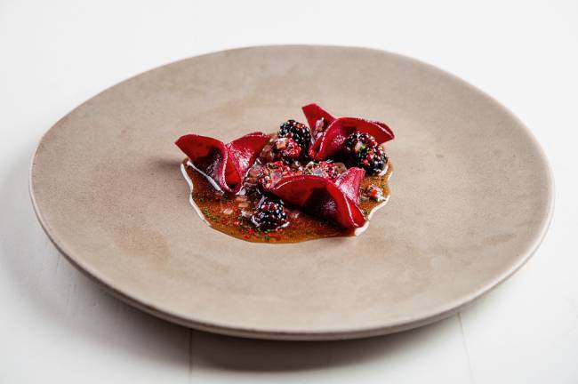 iFood Gourmet – Maní Ravióli de beterraba com molho de amora concentrado no centro de prato grande de cerâmica cinza.