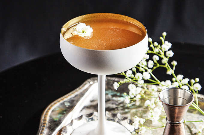 Foto do drinque banana daiquiri em taça metálica esmaltada de branco sobre bandeja de prata ao lado de ramo de florzinhas brancas.