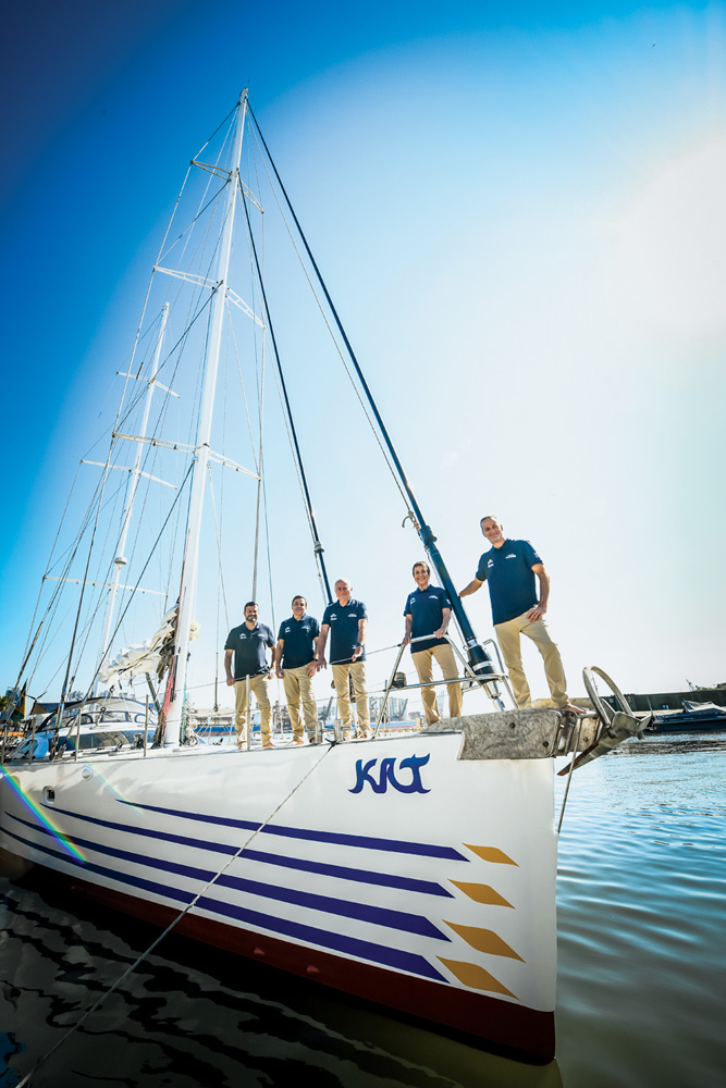 veleiro-kat-familia-schurmann-nova-aventura-poluicao-oceanos-historias-navegacao.JPG membros da família schurmann posando para a foto em fila, de lado, em cima da proa do barco kat. o fotógrafo está fora do barco para fazer a foto
