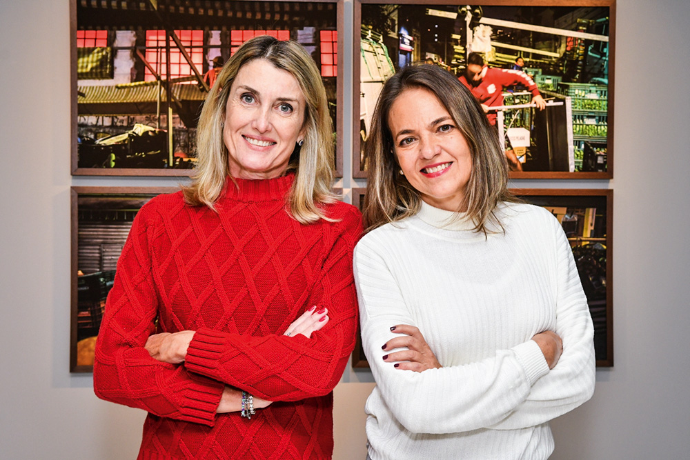 Dani Tranchesi e a sócia Paula Rocha aparecem lado a lado. Dani é loira e veste suéter vermelho. Paula tem cabelos castanhos e veste suéter branco.