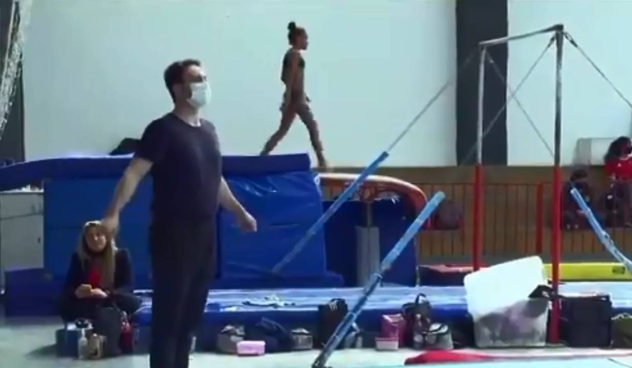 A imagem mostra Diego em um ginásio de Ginástica, em um grande quadrado de tatames, usado para a prática do solo. A imagem mostra ele de pé, com os braços estendidos, preparando para o salto.