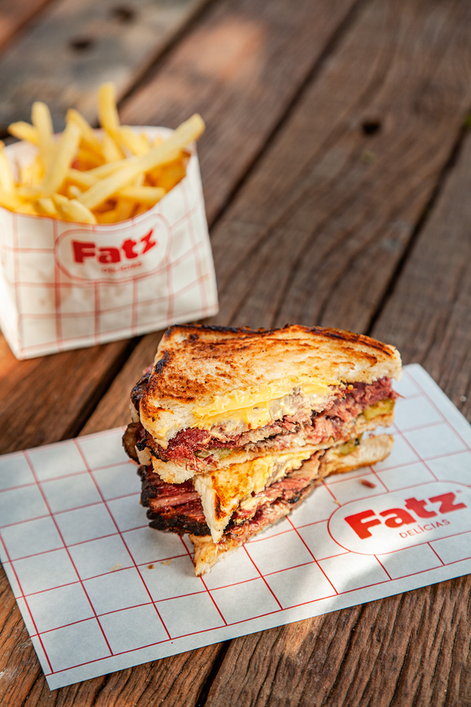 Fatz Delícias Dois pedaços, um sobre o outro, de sanduíches em pão de fôrma recheados de queijo cremoso e pastrami em cima de papel listrado branco e vermelho sobre mesa de madeira. Ao fundo batatas fritas em saquinho branco.