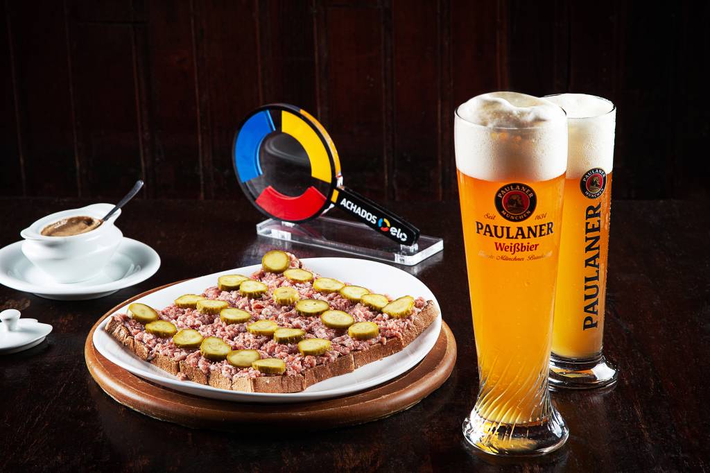 Achados ELO – Zur Alten Mühle Dois copos altos de chope com bastante colarinho à direita. Prato de louça branca sobre sousplat de madeira com canapés de carne com picles à esquerda.